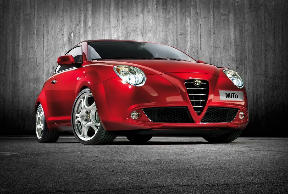 Um Alfa Romeo MiTo vermelho está estacionado em frente a uma parede de concreto texturizada. Os faróis estão acesos, refletindo em seu exterior brilhante e rodas de liga leve. Este estimado veículo não só exibe um design esportivo, mas também incorpora a renomada fiabilidade associada à marca Alfa Romeo. | Velocidade5.cv