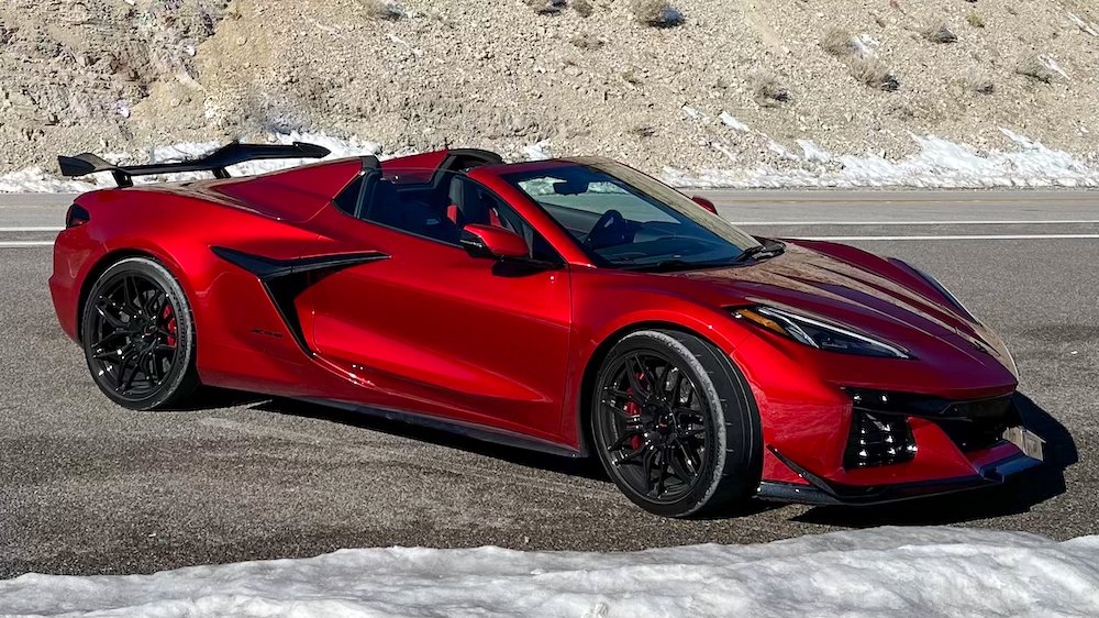 Um carro esportivo conversível Corvette Z06 vermelho está estacionado na lateral de uma estrada com neve. O carro apresenta um design elegante, rodas pretas e um spoiler traseiro proeminente, incorporando poder inigualável com seu motor V8. Uma estrada coberta de neve e terreno rochoso são visíveis ao fundo. | Velocidade5.cv