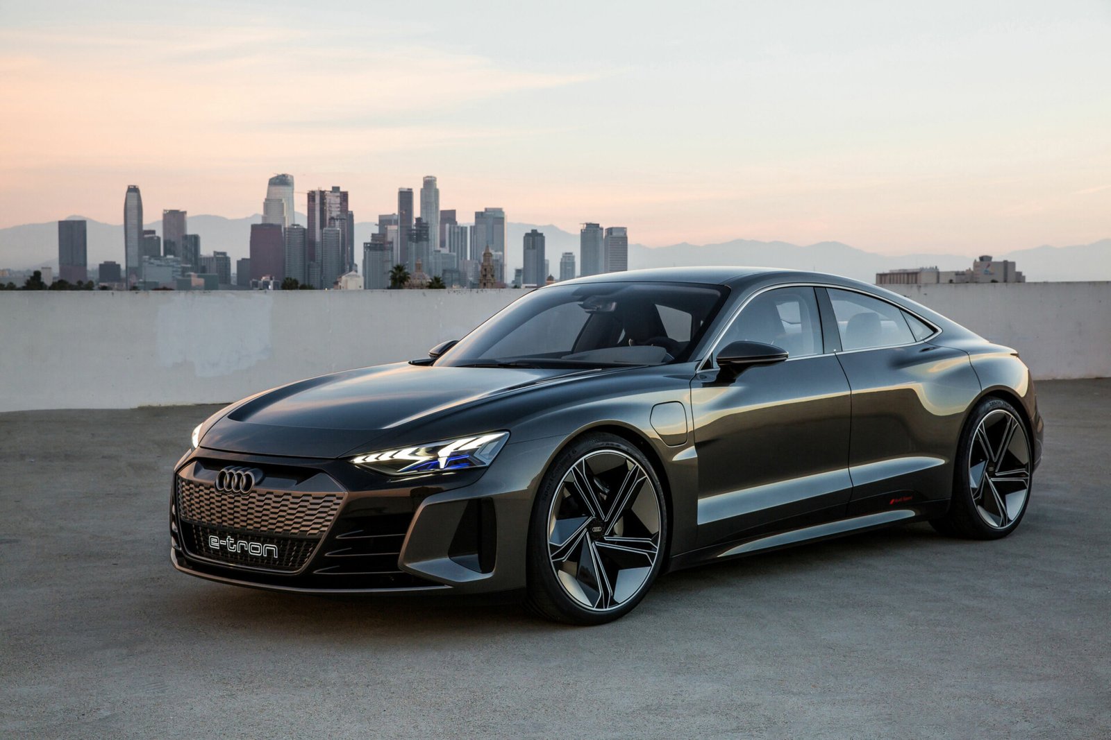 Um carro conceito Audi e-tron cinza escuro e elegante está estacionado em um telhado com um horizonte de cidade ao fundo. Como um dos melhores carros elétricos, ele apresenta faróis de LED distintos e curvas aerodinâmicas, sob um céu nublado com um sol poente ou nascente. | Velocidade5.cv