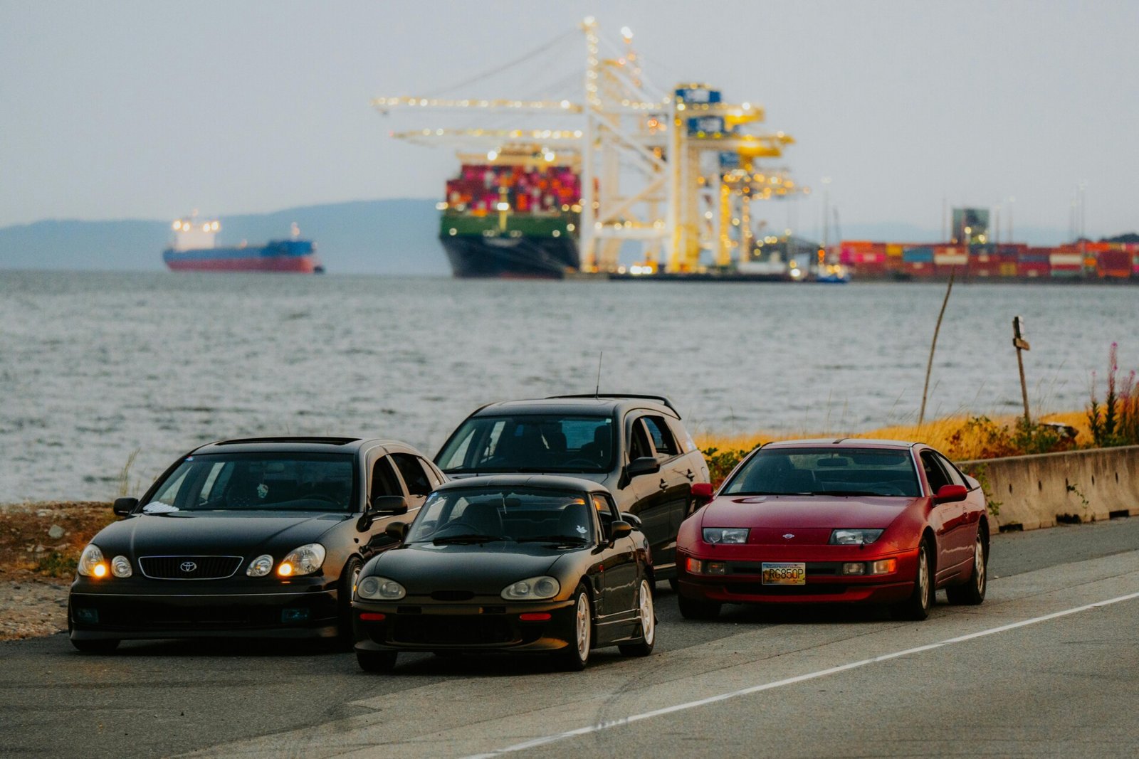 Três carros estacionados — um sedã preto, um SUV escuro e um carro esportivo vermelho — estão em uma estrada perto da água. Ao fundo, um grande navio de carga e guindastes industriais sugerem importação de carros neste cenário portuário. O céu parece nublado ou encoberto. | Velocidade5.cv