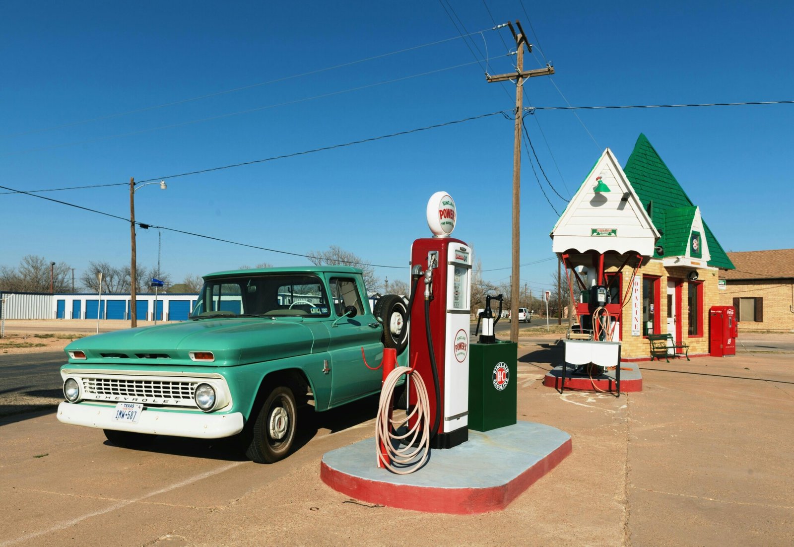 Uma caminhonete verde vintage está estacionada ao lado de um posto de gasolina antigo com um pequeno prédio com telhado verde. As bombas de gasolina em estilo retrô exibem o tipo de combustível disponível, e a cena se passa em um dia claro e ensolarado com linhas de energia ao fundo. | Velocidade5.cv