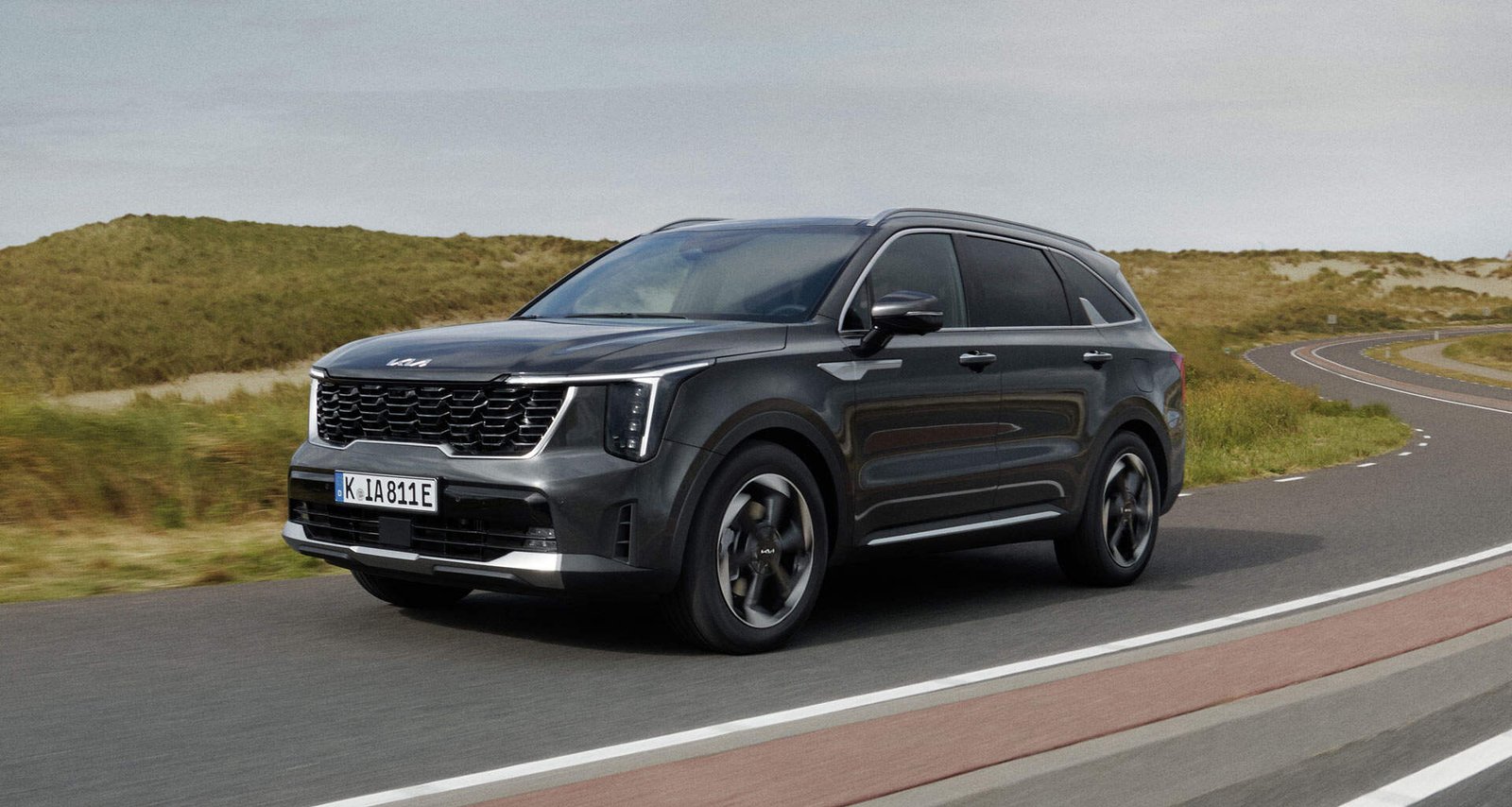 Um SUV híbrido de cor escura está dirigindo em uma estrada pavimentada cercada por dunas gramadas. O veículo está posicionado em um ângulo, mostrando sua lateral e frente em movimento enquanto navega pelo caminho. O céu está nublado, adicionando uma sensação de aventura a esta cena familiar. | Velocidade5.cv