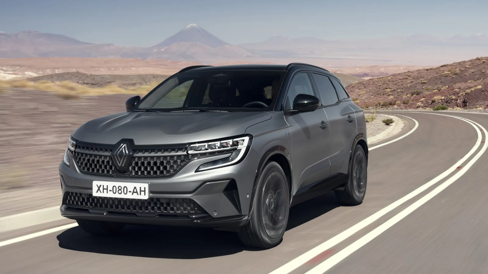 Um híbrido SUV Renault Austral cinza desliza por uma estrada sinuosa em uma paisagem desértica, ladeada por montanhas majestosas. O veículo ostenta um design elegante com faróis modernos e uma placa visível. Sob o céu limpo, este amigo do ambiente promete aventura em um cenário árido. | Velocidade5.cv