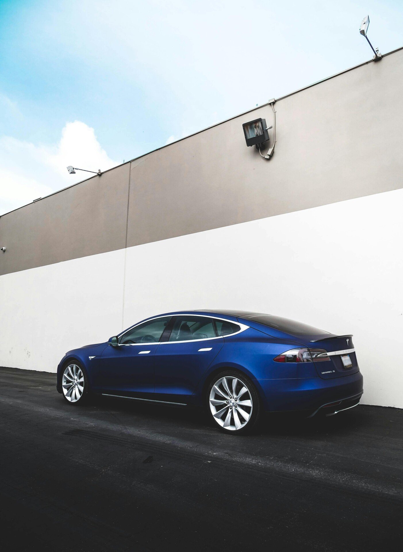 Um sedã elétrico azul com autonomia inigualável está estacionado ao lado de um grande muro de prédio bege e branco. O carro ostenta um design elegante com rodas de liga leve brilhantes, e o céu acima está limpo com algumas nuvens. | Velocidade5.cv