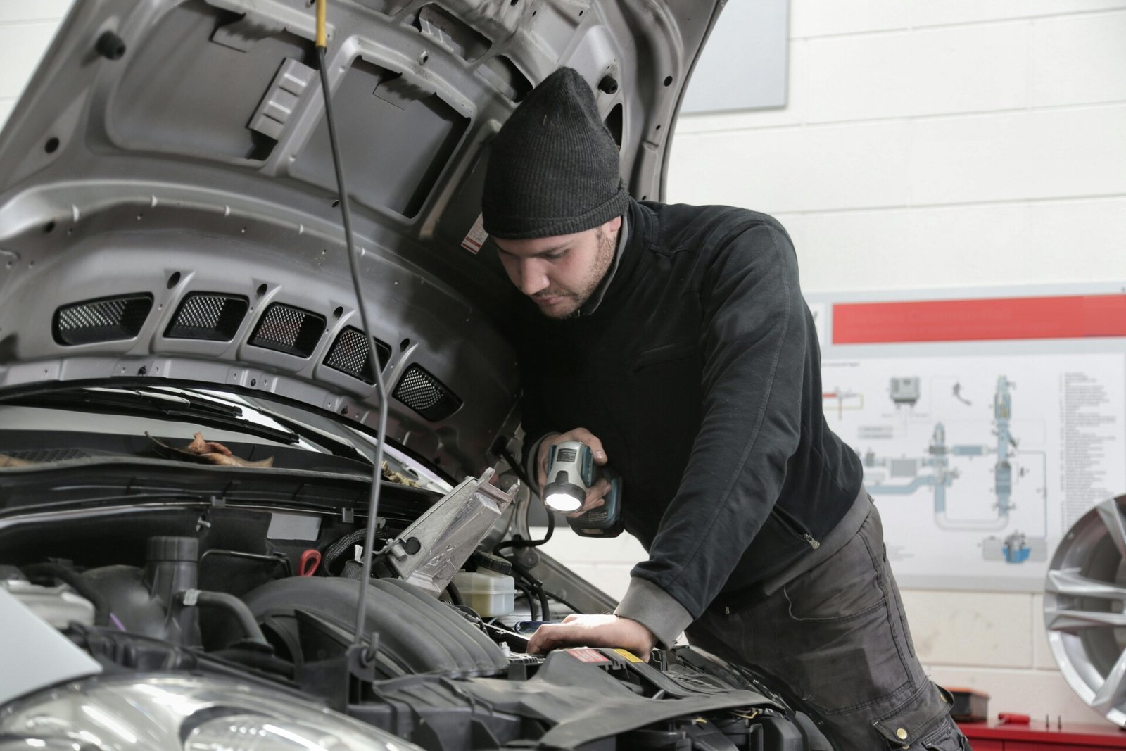 Um mecânico de gorro está inspecionando um motor de carro com uma luz portátil, garantindo que a correia dentada esteja em boas condições. O capô do carro está aberto, revelando seu funcionamento interno, enquanto diagramas adornam a parede ao fundo. Não adie manutenção necessária para desempenho ideal. | Velocidade5.cv