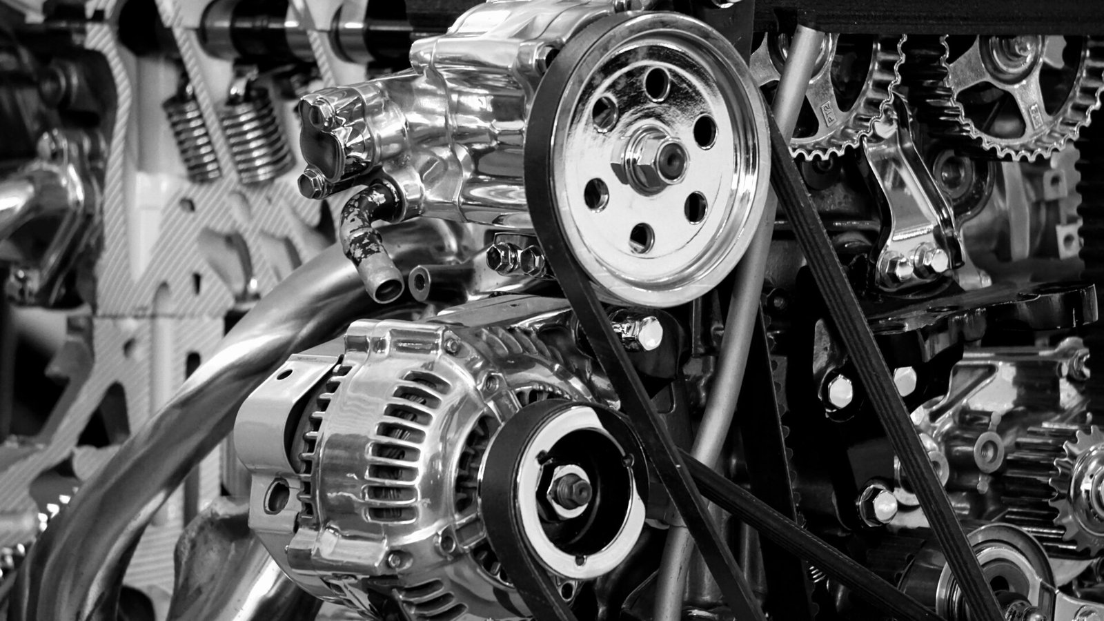 Visão de perto de um motor mecânico complexo e brilhante com engrenagens, correias e vários componentes visíveis. A imagem é em preto e branco, destacando as superfícies metálicas reflexivas e detalhes intrincados — um lembrete impressionante da precisão necessária para tarefas como a Mudança da Correia de Distribuição. | Velocidade5.cv